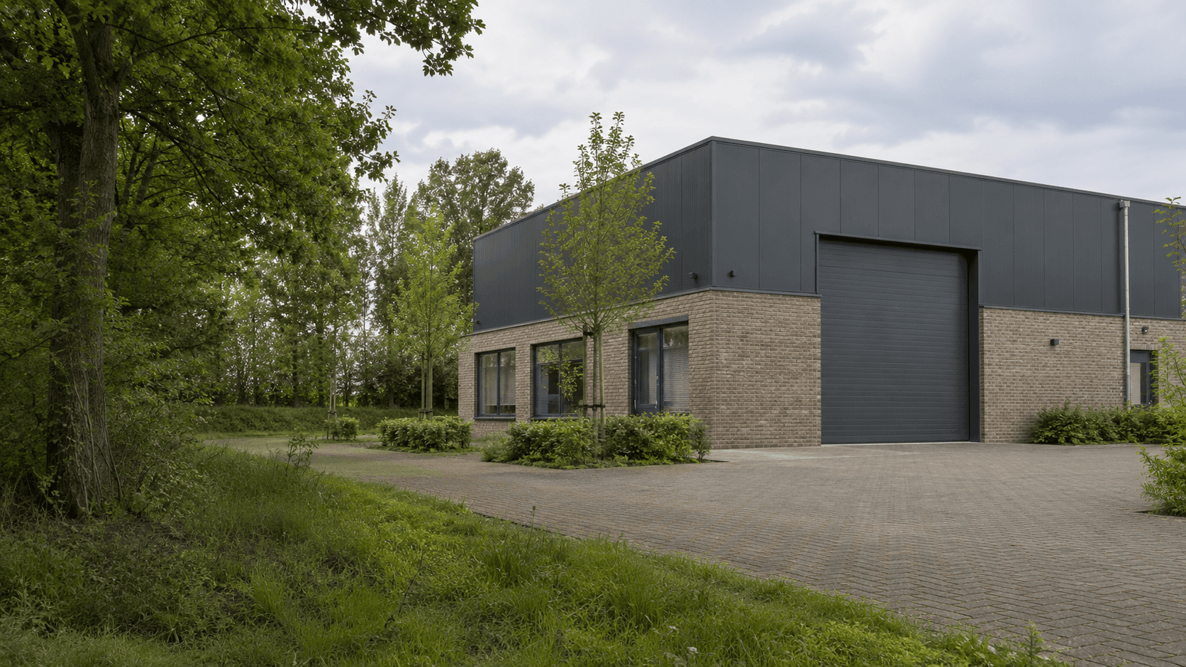 Bedrijfspand van baksteen en antraciet metaal met een gesloten rolpoort en jonge bomen langs het erf